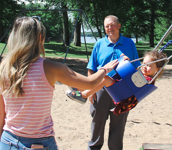 Scott Krug smiles nearby as a woman pushes a child in a blue swing at a sandy Wisconsin park.