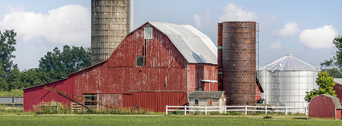 Weathered red barn with silos and a white fence in rural Wisconsin under a blue sky.
