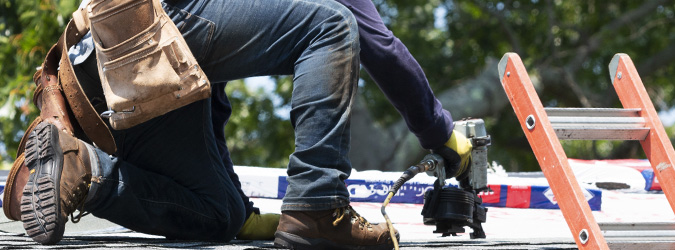 A construction worker kneels on a roof with tools, near a ladder and Wisconsin building materials.